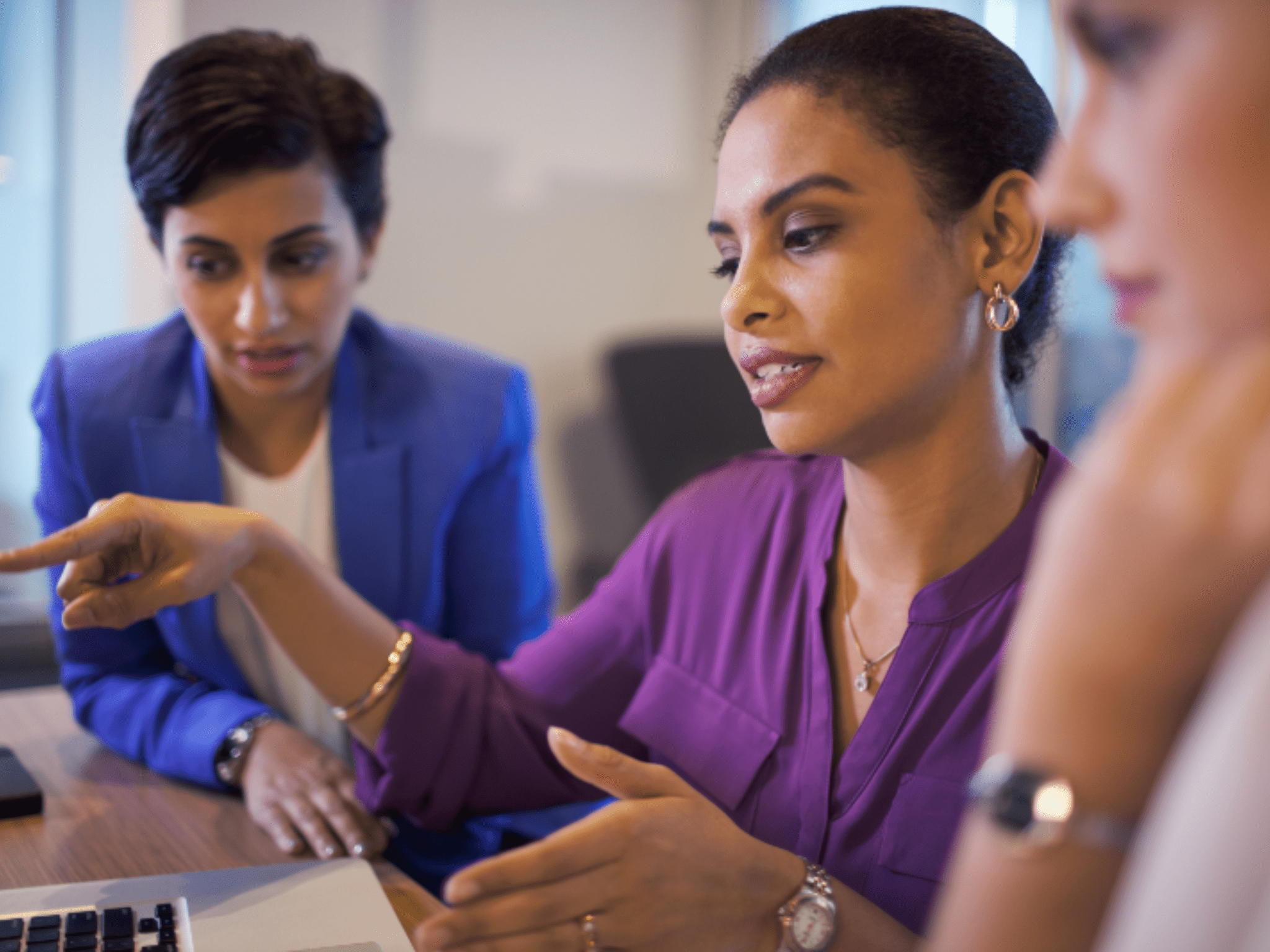 A women pointing to a computer monitor offscreen while 2 women look on.