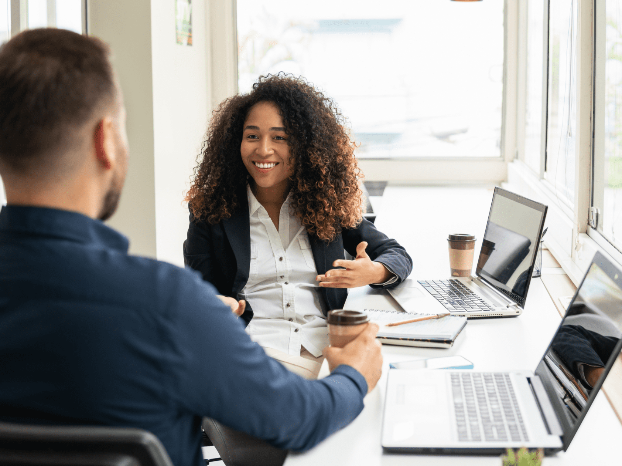 Woman smiling and gesturing to someone while they are both seated in front of laptops at shared desk.