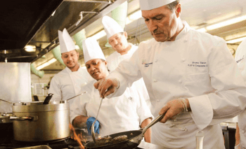 Student chefs learning by watching the head chef cook at a stove