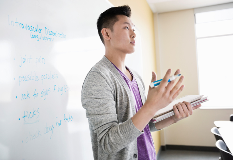 A young man holding a dry erase marker and notebook standing in front of a whiteboard